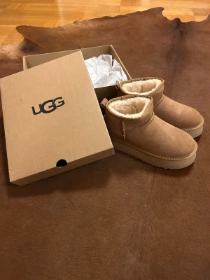 Ugg - Säljer ett par UGG boots i beige mocka med mjukt, fluffigt foder och plattformsula. Skorna har rund tå och grov yttersula för extra grepp. Klassisk UGG-logga baktill och dragflik för enkel påtagning. Perfekta för kalla dagar och riktigt cozy vibes.