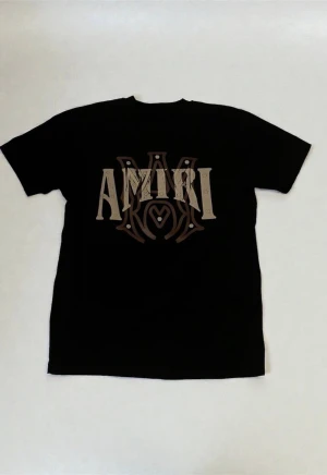 AMIRI t-shirt - Svart t-shirt från AMIRI i bomull med stor beige och brun logotyp framtill. Klassisk rund hals och korta ärmar. Snyggt tryck som ger en cool vibe och passar perfekt till streetwear-stilen. Köpt från farfetch.