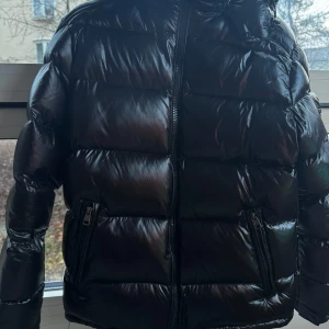 Svart Moncler pufferjacka med huva - Säljer en svart pufferjacka från Moncler med glansig finish och huva. Jackan har två fickor med dragkedja och klassisk Moncler-logga på ärmen. Perfekt för dig som vill ha en snygg och varm jacka med street vibe.