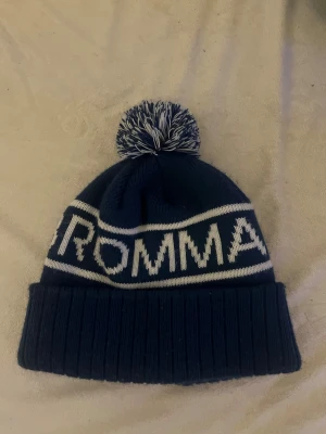 Bromma mössa - Säljer en mörkblå Bromma-mössa med vit text och en fluffig tofs i blått och vitt. Tjockt och varmt stickat tyg som är perfekt för kalla dagar. En snygg och sportig mössa som håller huvudet varmt samtidigt som du visar vilket lag/ställe du supportar.