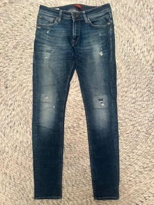 Jack & Jones Skinny Liam jeans blå - Snygga blå skinny jeans från Jack & Jones, modell Liam. Jeansen har slitna detaljer och lagningar framtill, klassiska fem fickor och läderpatch bak i midjan. De är tillverkade i stretchig denim för en tight passform och har en modern, smal siluett.