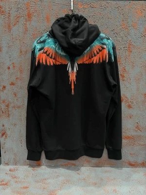 Svart hoodie County of Milan med vingar - Svart hoodie från County of Milan med fett tryck av färgglada vingar i orange, turkos och vitt över axlar och bröst. Tröjan har huva med snörning och stor magficka framtill. Perfekt för dig som vill sticka ut med en unik streetstyle.