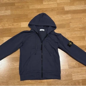 Mörkblå hoodie från Stone Island - Snygg mörkblå hoodie från Stone Island med klassisk logotyp-patch på vänster ärm. Tröjan har huva med snörning, dragkedja framtill och två fickor. Tillverkad i mjuk bomull för skön komfort och stilren look. Perfekt för dig som gillar streetwear och vill sticka ut.