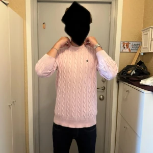 Ljusrosa kabelstickad tröja Morris - Säljer en ljusrosa kabelstickad tröja från Morris i 100% bomull. Tröjan har rund hals, ribbade muddar och en liten broderad logga på bröstet. Lite urtvättad där av priset 