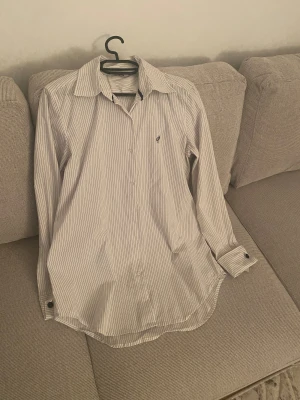 Vit randig skjorta från Gina, strl 42 - Snygg vit skjorta med tunna svarta ränder från Gina. Klassisk krage med svarta detaljer, långärmad och med knappar framtill. Broderad logga på bröstet. Perfekt för dig som gillar stilrena och tidlösa plagg.