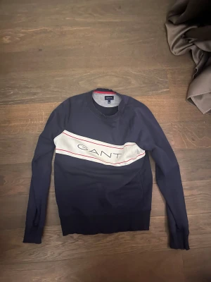 Mörkblå GANT tröja med logga - Snygg mörkblå tröja från GANT i storlek S. Tröjan har en vit panel med röd kant och stor GANT-logga över bröstet. Rund hals och långa ärmar med ribbade muddar. Perfekt för dig som gillar klassisk och sportig stil.