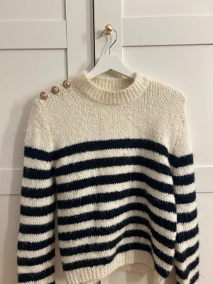 Randig stickad tröja från Zara - Mysig stickad tröja från Zara i storlek S. Tröjan är vit med mörkblå horisontella ränder och har tre dekorativa guldfärgade knappar på ena axeln. Rund hals och långa ärmar, perfekt för kyliga dagar.