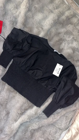 Svart blus med ballongärmar från NA-KD - Säljer en svart blus från NA-KD med ballongärmar och bred smock vid midjan och ärmsluten. Toppen har en croppad passform och är tillverkad i ett lätt syntetmaterial. Perfekt för dig som gillar trendiga detaljer och volym på ärmarna.