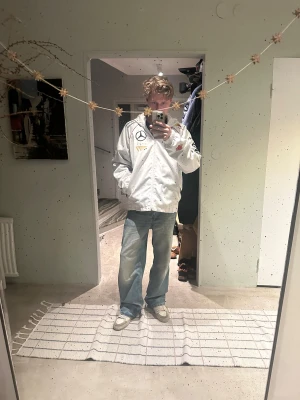 Vit vindjacka T1 x Mercedes Zeus - Säljer en vit vindjacka med huva från GOALSTUDIO x T1, med feta Mercedes, Red Bull och Samsung Odyssey-loggor samt ZEUS-tryck på ryggen. Jackan har dragkedja, svarta detaljer på sidorna och röda sömmar. Insidan har coolt mönstrat foder. 