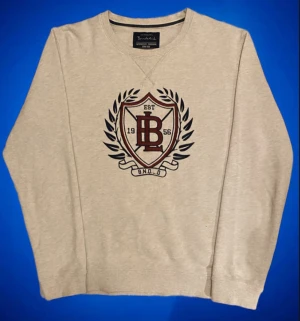 Bondelid Sweatshirt Grå - Snygg beige sweatshirt från Bondelid med stort broderat emblem i vinrött och marinblått på bröstet. Tröjan har rund hals, ribbade muddar och lång ärm. Perfekt för dig som gillar klassisk college-stil och vill ha en bekväm tröja med cool detalj framtill. Den är helt felfri sitter som S/M priset kan alltid diskuteras!👍