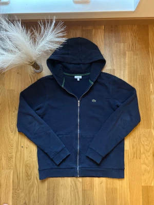 Marinblå hoodie från Lacoste - Säljer en marinblå hoodie från Lacoste med dragkedja och klassisk krokodillogo på.Tillverkad i mjuk bomulls- och polyestermix, perfekt för chill dagar. Ordinarie pris 1199kr, köpt ifrån kidsbrandstore. Pris kan diskuteras👌