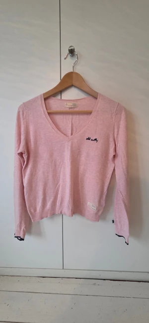 Rosa v-ringad tröja Odd Molly - Söt ljusrosa tröja från Odd Molly med v-ringning och lång ärm. Tröjan har en liten mörkblå rosett på ärmsluten och mörkblå broderad logga på bröstet. Materialet är mjukt och tunt, 100% bomull.. Svarta detaljer vid muddar ger en extra touch.