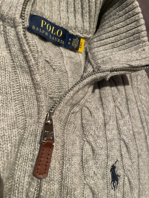 Kabelstickad half zip tröja Polo Ralph Lauren - Snygg grå stickad tröja från Polo Ralph Lauren med klassiskt kabelmönster och half zip med läderdetalj. Tröjan har ribbad krage, muddar och nederkant samt broderad logga på bröstet. Perfekt för dig som gillar stilren och tidlös design. Storlek M men passar även till S.