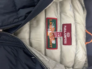 Svart dunjacka Parajumpers Y/M - Svart super lightweight dunjacka från Parajumpers med quiltad design och dragkedja framtill. Jackan har ståkrage, två sidofickor med dragkedja och en gul detalj vid halsen. Insidan är ljusbeige och materialet är syntet med dunfyllning.