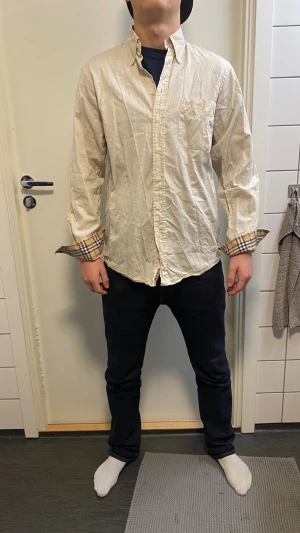 Beige Burberry skjorta med rutiga detaljer - Säljer en beige skjorta från Burberry med klassiska rutiga detaljer på insidan av manschetterna och kragen. Skjortan har lång ärm, bröstficka och knäppning framtill. Tillverkad i bomull och har en clean, tidlös look.