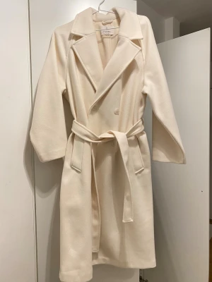 Vit kappa från Stradivarius XL - Snygg vit kappa från Stradivarius i storlek XL. Jackan har klassisk krage, knappar framtill och ett matchande bälte i midjan. Långärmad och med stilren design, perfekt för dig som gillar minimalistiskt och cleant. Materialet är mjukt och känns lyxigt.