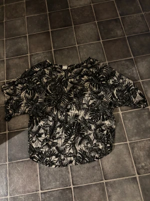 Svartvit bladmönstrad skjorta Gina Tricot - Snygg svartvit skjorta från Gina Tricot i storlek 40. Skjortan har ett coolt bladmönster, lös passform och korta ärmar. Perfekt för dig som gillar statement-plagg med tropisk vibe.