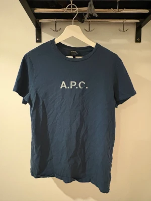 Blå t-shirt från A.P.C. med tryck - Snygg blå t-shirt från A.P.C. med klassiskt vitt logotryck på bröstet. Modellen är rak med rund hals och korta ärmar. Tillverkad i mjuk bomull som känns skön mot huden. Perfekt för dig som gillar stilrena och enkla plagg med en touch av Paris. Nypris: 1300kr