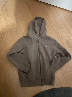 Brun hoodie från Polo Ralph Lauren - Snygg brun hoodie från Polo Ralph Lauren med dragkedja och klassisk vit logga på bröstet. Modellen har huva, känguruficka och ribbade muddar. Tillverkad i mjuk bomullsblandning som är skön att ha på sig.