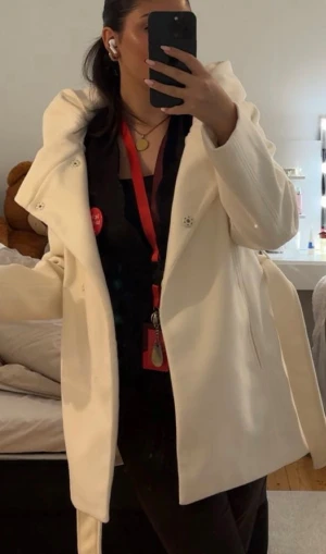 Vit kappa med luva  - Snygg vit kappa med bred luva och knappar framtill. Jackan har en rak passform och är tillverkad i ett mjukt tyg som känns lyxigt. Perfekt för dig som vill ha en stilren och clean look under kallare dagar. Den är varm och använd endast en gång, köpt för 799kr