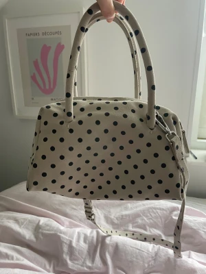 Prickig handväska från Zara - Snygg handväska från Zara i vit mocka med svarta prickar. Väskan har två handtag, avtagbar axelrem och dragkedja upptill. Insidan är svart med en innerficka med dragkedja. Perfekt storlek för det viktigaste och sticker ut med sitt coola prickiga mönster. Aldrig använd och köpt för 999kr.