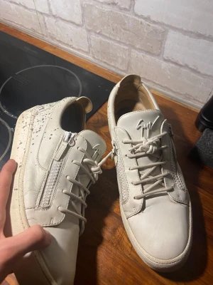 Giuseppe Zanotti vita sneakers strl 45 - Säljer ett par vita sneakers från Giuseppe Zanotti i storlek 45. Skorna har snörning, dragkedjor på sidorna och en rund tå. Tillverkade i skinn med en platt sula och signatur-logga på plösen. Snygg italiensk design med stilrena detaljer.