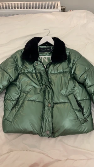 Glansig jacka Calvin Klein - Snygg grön pufferjacka från Calvin Klein i storlek xs men den är lite större i modellen (s). Köptes i USA och är inte tillgänglig i Sverige! Svart fuskpäls på kragen. Jackan har dragkedja och knappar framtill, två sidofickor med dragkedja och en innerficka. Perfekt för vintern! Hyfsat ny (har bara haft i 6månader) men säljer för att den inte kommer till användning! Skriv för mer bilder! Priset kan diskuteras