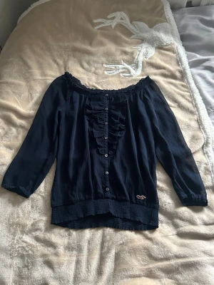 Mörkblå offshoulder blus Hollister XS - Snygg mörkblå offshoulder blus från Hollister i tunn, plisserad polyester. Blusen har trekvartsärm, knappar framtill och volangdetaljer längs knappraden. Smockad kant upptill och nertill, samt Hollister-logga broderad vid nederkanten. Perfekt för dig som gillar en chill och feminin stil.