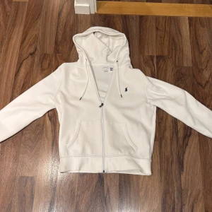Vit hoodie från Polo Ralph Lauren - Snygg vit hoodie från Polo Ralph Lauren med dragkedja, huva med dragsko och klassisk broderad logga på bröstet. Mjuk bomullsblandning och två stora fickor framtill. Perfekt för en clean och stilren look.