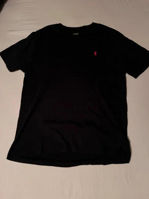 Svart t-shirt Polo Ralph Lauren XL - Svart t-shirt från Polo Ralph Lauren i storlek XL. Klassisk modell med korta ärmar och rund hals. Ikoniska röda Polo-loggan broderad på bröstet. Tillverkad i mjuk bomull för en skön känsla. Perfekt basic med stilren vibe.