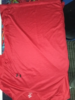 Röd Under Armour t-shirt i polyester - Snygg röd t-shirt från Under Armour med svart logga på bröstet och ryggen. Tillverkad i mjuk och lätt polyester som andas, perfekt för träning. Klassisk rund hals och korta ärmar, passar dig som gillar sportig stil.