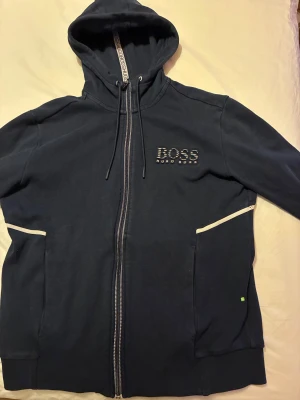 Mörkblå hoodie från Hugo Boss - Snygg mörkblå hoodie från Hugo Boss med dragkedja och huva med snörning. Stora fickor framtill och vita detaljer längs fickorna. Boss-logga broderad på bröstet och tryck i luvan. Mjuk bomullskänsla, perfekt för chill dagar.