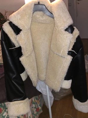 Svart teddyjacka med fuskpäls - Säljer en svart teddyjacka med krage och detaljer i beige fuskpäls. Jackan har breda slag, synliga sömmar och är fodrad med mjukt teddyfoder. Utsidan är i svart syntetmaterial som ger en skinnlook, medan insidan och muddar är fluffiga och varma.