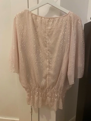 Glittrig champagne/ljusrosa blus - Säljer en superfin ljusrosa blus med glitter i guld. Blusen har puffig passform, korta ärmar och bred volang nedtill med smock. Materialet är lätt och luftigt, perfekt för fest. Rund hals med liten v-ringad detalj. 