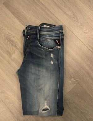 Replay Anbass blå jeans med slitningar - Snygga Replay Anbass jeans i blå tvätt med slitna detaljer och hål på framsidan. Klassisk femficksmodell med raka ben och normal passform. Tydliga Replay-loggor på ficka och bakre läderpatch. Perfekt för dig som gillar en cool och avslappnad stil. Storleken är 31/32 men anpassas till din kropp pågrund av att dem är hyperflex 