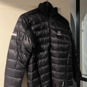 Svart dunjacka Haglöfs XS - Svart dunjacka från Haglöfs i storlek XS. Jackan har ett stilrent, quiltat mönster och hög krage. Materialet är glansigt och lätt, perfekt för kalla dagar. Diskret logga på bröstet och ärmen. Dragkedja framtill och långärmad modell.
