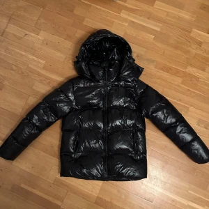 Svart pufferjacka - Snygg svart pufferjacka från Everest med glansig finish och huva. Jackan har dragkedja framtill, två fickor med dragkedja och en klassisk badge på ärmen. Perfekt för kalla dagar när du vill hålla stilen och värmen.