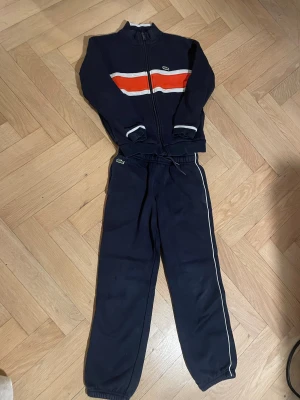 Tränings sett från Lacoste - Säljer en mörkblå träningjackafrån Lacoste med dragkedja framtill och coola röda och vita ränder över bröstet. Det ingår också dom mörkblåa Lacoste mjukis byxorna. Storlek 8år
