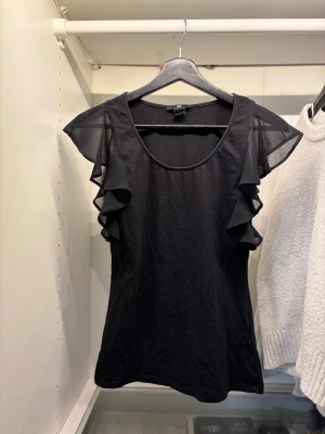 Svart topp med volangärmar från H&M - Svart topp från H&M med transparenta volangärmar i mesh. Rund halsringning och figurnära passform. Perfekt för dig som gillar en enkel men ändå cool look. Storlek xs/s
