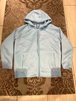 Ljusblå Nike NOCTA hoodiejacka - Säljer en ljusblå hoodiejacka från Nike NOCTA med dragkedja hela vägen, två fickor med dragkedja och huva. Jackan har en clean look med NOCTA-logga i nacken och Nike Swoosh på bröstet. Perfekt för dig som gillar streetwear och sportig stil.