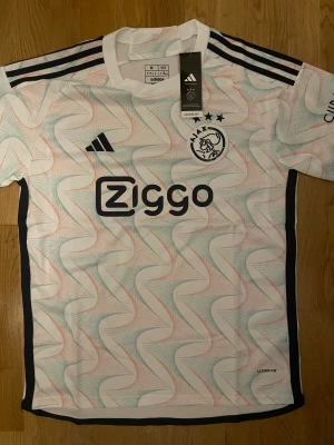 Ajax x Adidas vit mönstrad fotbollströja - Ajax fotbollströja från Adidas i slim fit med korta ärmar. Tröjan är vit med grafiskt mönster i ljusblått och ljusrött, har svarta detaljer och Ziggo-logga på bröstet. Tillverkad i lätt polyester med Aeroready-teknologi för extra komfort. Perfekt för dig som älskar fotboll och snygg design.