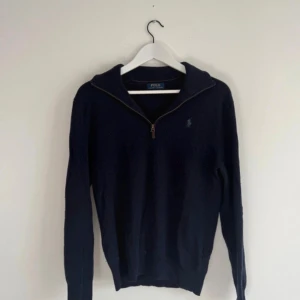 Mörkblå halvziptröja Polo Ralph Lauren - Mörkblå stickad tröja från Polo Ralph Lauren med halv dragkedja och klassisk krage. Tröjan har lång ärm och diskret broderad logga på bröstet. Perfekt för dig som gillar stilren och tidlös design.