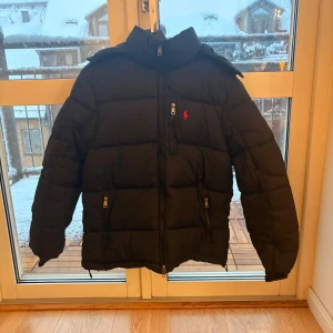 Polo Ralph Lauren Vinterjacka  - Svart dunjacka från Polo Ralph Lauren i storlek M. Jackan har en hög krage, huva och flera dragkedjefickor. Klassisk pufferstil med röd logga på bröstet. Perfekt för kalla dagar och har en snygg, stilren look. Kvitto finns 💞🥰