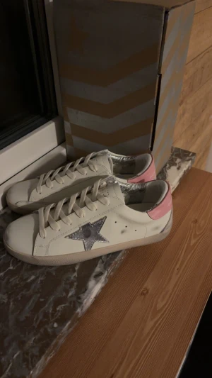 Golden Goose vita sneakers med rosa detalj - Golden Goose sneakers i vitt skinn med rosa hälparti och silverfärgad stjärna på sidan. Skorna har snörning, rund tå och platt sula. Insidan är fodrad med silverdetaljer och yttersulan är svart. Perfekt för dig som gillar coola och trendiga sneakers.