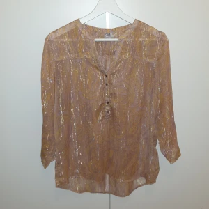 Glittrig blus från Saint Tropez - Glittrig blus från Saint Tropez i storlek S. Blusen är aprikos med guldiga inslag och har ett paisleymönster samt knappar framtill. Den är tunn och luftig med trekvartsärm och har en lätt transparent känsla. 