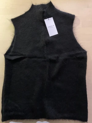 Svart stickad väst från Stockh LM Studio - Svart, mjuk och fluffig stickad väst från Stockh LM Studio i storlek M. Västen har hög krage och är tillverkad i ett lurvigt material som ger en cool och mysig vibe. Perfekt att styla över en långärmad tröja för extra edge.