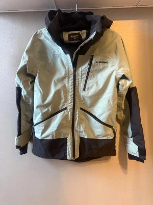 Everest skidjacka  med huva .stl 152 - Snygg skidjacka från Everest i svart och ljusgrått med flera dragkedjefickor och justerbara ärmslut med kardborre. Jackan har huva och coola kontrastpaneler, perfekt för dig som gillar outdoor och streetstyle. Materialet är slitstark polyester.