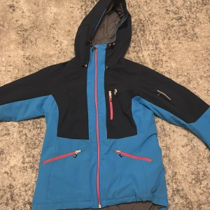 Blå och svart skidjacka Peak Performance - Peak Performance skidjacka i blå med huva och rosa dragkedjor. Jackan har flera fickor + skidkortsficka på vänsterarmen.