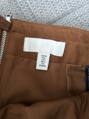 Brun kortkjol från H&M i mocka-look - Snygg brun kortkjol från H&M i storlek 32. Kjolen har en rak passform, dragkedja bak och två dekorativa fickor. Materialet har en mocka-liknande känsla som ger en trendig vibe. Perfekt att styla med stickad tröja eller t-shirt.