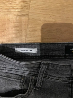 Grå slim jeans från Jack & Jones - Snygga grå jeans från Jack & Jones i modellen Slim/Glenn. De har en smal passform, klassisk femficksdesign och svarta detaljer på bakfickan. Jeansen är tillverkade i mjukt denimtyg med lätt tvättad look och passar perfekt till sneakers eller boots.